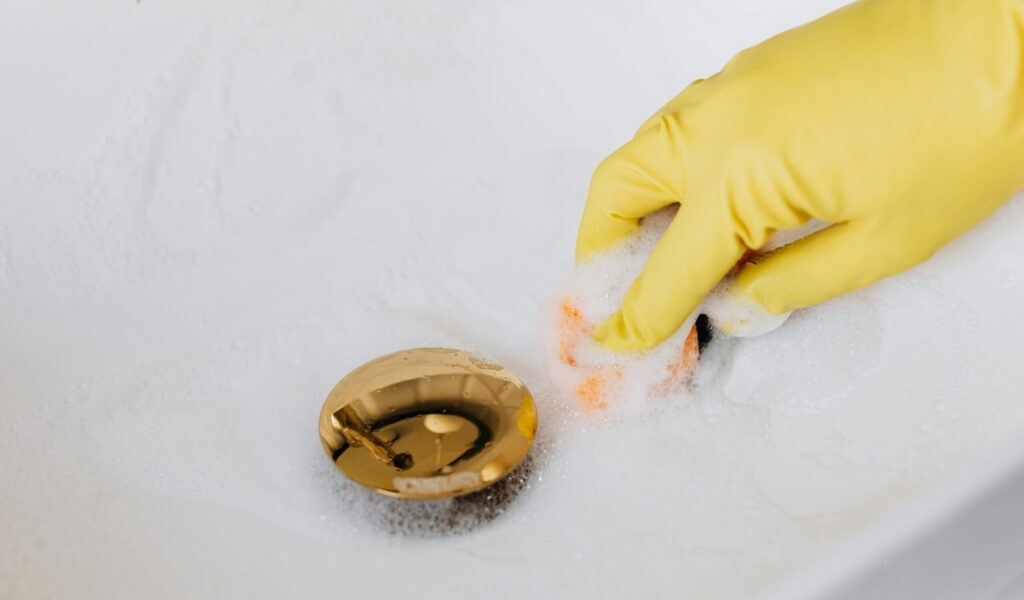 From above of crop anonymous person in yellow rubber protective glove using sponge and detergent for washing white sink in bathroom